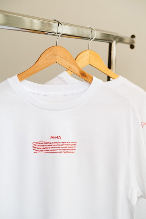 Dos camisetas blancas con texto rojo frontal de GEN ES colgadas de una percha
